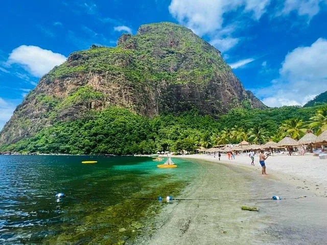 A large green mountain looms over a sandy beach with thatched umbrellas.