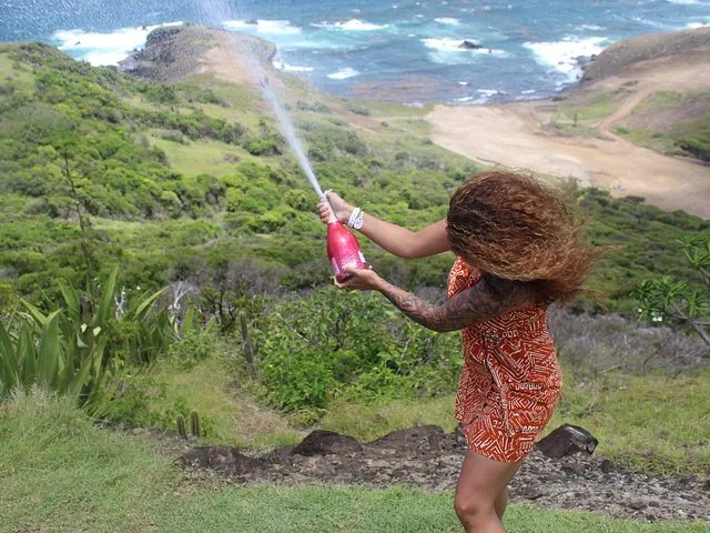 A woman popping a bottle of champagne on a grassy hillside overlooking the ocean.