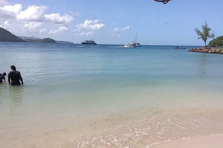 People wading in shallow beach water with boats anchored in the distance.
