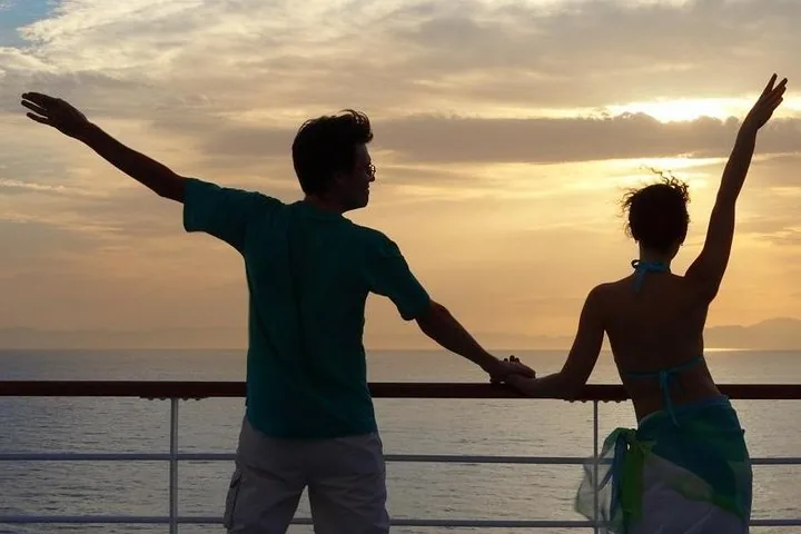 Silhouetted couple standing on a boat railing with arms raised at sunset.